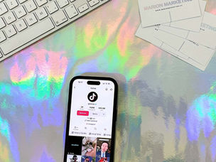 Mobilní telefon leží na duhovém pozadí vedle vizitek Marion marketing a bílé klávesnice. TikTok reklama cena – a jak stanovit rozpočet