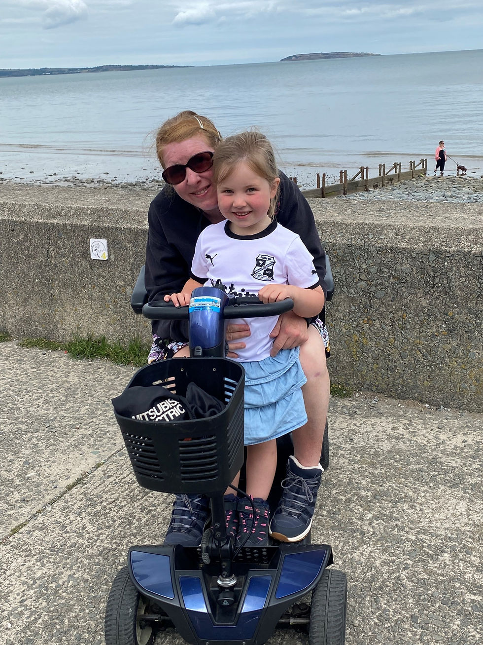 Woman with girl on her lap, sitting on a mobility scooter by the seaside. Both smiling. Cloudy sky, calm water, person walking dog in background.