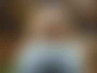 A person with wavy blonde hair wearing a hooded sweatshirt adorned with a cross symbol explores a serene forest path amidst the colors of autumn.