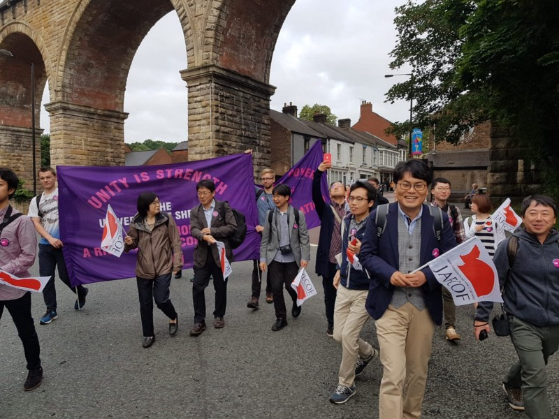 A group of people carrying flags and banners walking past a railway bridge