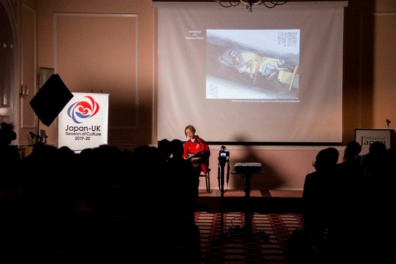 A woman reading in front of a screen with an image of mining in the background