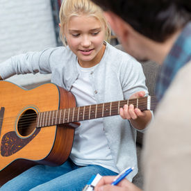Young guitar student smiling during a music lesson, holding her guitar while her teacher listens attentively with a notepad in hand at Kingwood Arts Academy of Music, Kingwood, Tx.