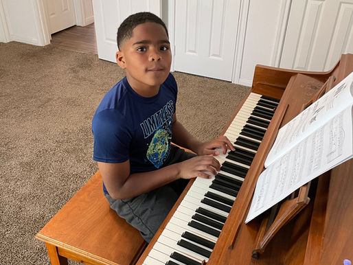 A young piano student practicing at an upright piano with sheet music open, developing beginner keyboard skills and confidence during a piano lesson at Kingwood Arts Academy of Music, in Kingwood, Texas.