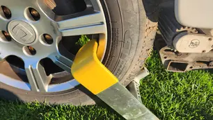 Car tire with yellow wheel clamp, parked on green grass. Visible car wheel details, no other text or people present.