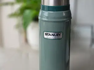 Green Stanley thermos with silver lid on a white surface, blurred plant in background. Text: "STANLEY SINCE 1913."