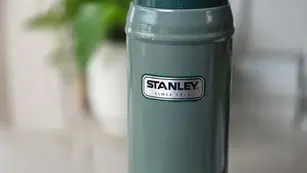 Green Stanley thermos with silver lid on a white surface, blurred plant in background. Text: "STANLEY SINCE 1913."
