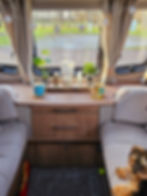 Cosy caravan interior with plants, candles, and cups on a table. A small dog sits on a grey sofa. Outside view of trees and grass. Relaxed vibe.