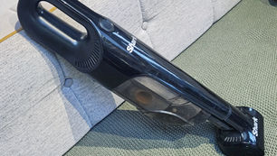 Black handheld Shark vacuum leaning on a light gray couch, set on a green carpet. The vacuum is sleek and modern, showcasing its compact design.