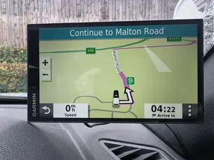 Garmin GPS screen in a car shows a navigation route: "Continue to Malton Road." Speed is 0 mph, arrival in 4:22. Background shows a wooden fence.