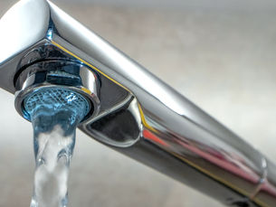 Shiny silver faucet with water pouring out against a blurred beige background. Reflection visible on the metal surface.