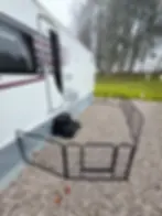 Caravan with black and red stripes parked on gravel. Black wire pen enclosing area. Leaf litter on ground; misty trees in background.
