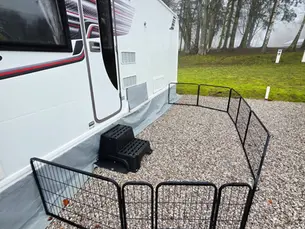 Caravan with black and red stripes parked on gravel. Black wire pen enclosing area. Leaf litter on ground; misty trees in background.