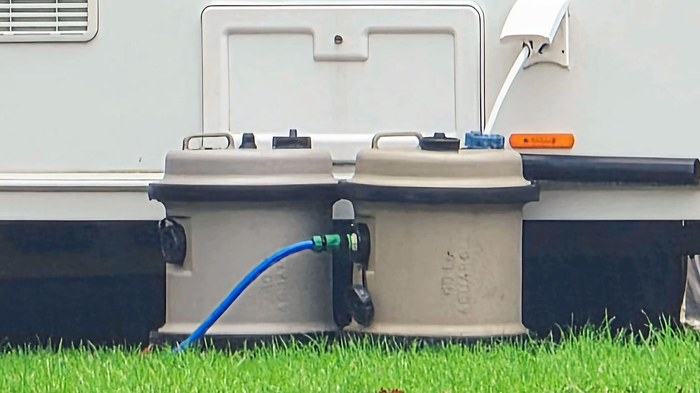 Two gray aquaroll water carriers connected by a blue hose sit on green grass, adjacent to a white caravan. The setting appears calm and outdoors.