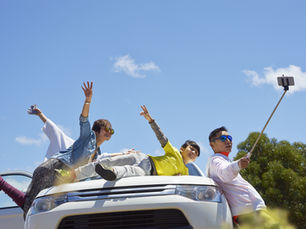 Three people on a car hood, excitedly posing for a selfie on a sunny day. Clear blue sky, trees in background, joyful mood.