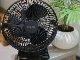 Black tabletop fan labeled "Koonie" near a green potted plant on a beige surface. Background suggests a cosy indoor setting.