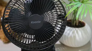 Black tabletop fan labeled "Koonie" near a green potted plant on a beige surface. Background suggests a cosy indoor setting.