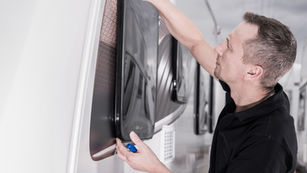 Man adjusts a caravan window on a white wall, holding a blue tool. Indoor setting with a focused expression.