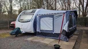 White caravan with a grey awning parked on gravel near trees. A red car is partially visible, and a small potted plant is in front.
