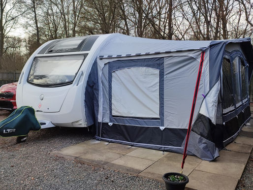 White caravan with a grey awning parked on gravel near trees. A red car is partially visible, and a small potted plant is in front.