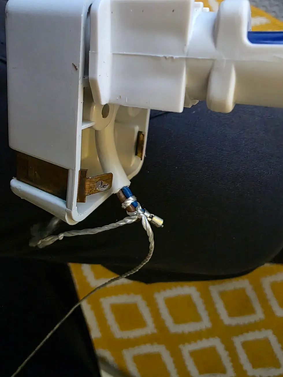 White plastic electrical plug with exposed wires rests on dark pants. Background shows yellow and white geometric pattern.