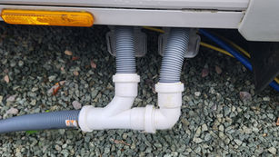 Gray drainage hoses connected to white pipes under a vehicle, set against a gravel background with visible yellow reflector.