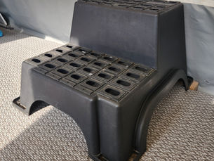 A black plastic step stool with perforated top is on a textured carpet in front of a white caravan wall, lit by natural light.
