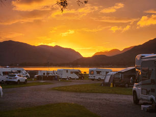 Campers and cars parked by a serene lake at sunset. Mountains silhouette the horizon, with a vibrant orange sky setting a peaceful mood.