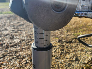 Close-up of a metal hitch with a weight gauge marked in kgs on a sunny day. Background shows blurred gravel and grass.