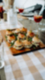 Mini sliders on a wooden board with pickles and arugula, set on a checkered tablecloth. Glasses with straws in the background.