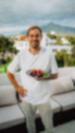 Smiling chef in white holds a platter of skewered treats with raspberries. Outdoor setting with mountains and cloudy sky in the background.