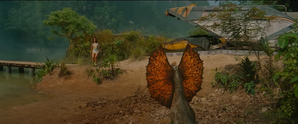 ฉากตื่นเต้นจากภาพยนตร์ Jurassic World: Rebirth ถ่ายทำที่นายเหมือง ตำบลทับปริก จังหวัดกระบี่ แสดงภาพไดโลโฟซอรัสเผชิญหน้ากับหญิงสาวริมฝั่งน้ำ ข้างยานพาหนะล้ำยุค ท่ามกลางฉากหลังป่าร้อนชื้น