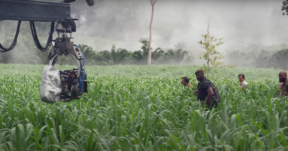 ภาพเบื้องหลังการถ่ายทำภาพยนตร์ Jurassic World: Rebirth ที่สวนปาล์ม ตำบลเขาคราม จังหวัดกระบี่ นักแสดงเดินผ่านทุ่งหญ้าเขียวขจี ขณะกล้องเครนกำลังบันทึกภาพ ท่ามกลางฉากหลังป่าร้อนชื้นและหมอกจาง
