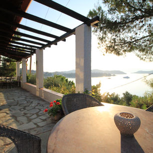 Im Vordergrund der rote runde Marmortisch auf der großen Terasse. Im Hintergrund der Sonnenaufgang über der Bucht von Megali Ammos