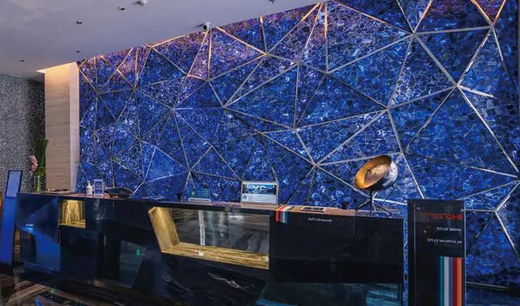 Eine glänzend schwarze Empfangstheke mit goldenem Einsatz steht vor einer blauen Steinwand mit feinen geometrischen Linien.