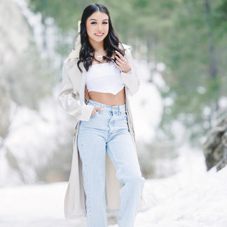 Jasmin standing confidently in the snow wearing a white crop top, jeans, and a beige coat, with a bright forest backdrop.
