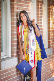 Diana leaning against a brick wall confidently in front of Old Main, proudly showcasing her graduation stole and honors cords.