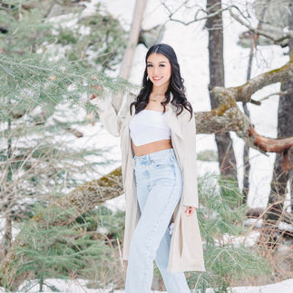 Jasmin standing amidst snow-covered trees, dressed casually in a beige coat and jeans, holding a pine branch gently