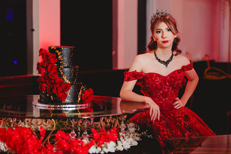 Quinceanera standing beside her luxurious black and gold cake adorned with red roses.