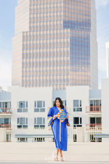 Graduate in a royal blue gown standing on a rooftop, holding her decorated cap with a city skyline behind her.