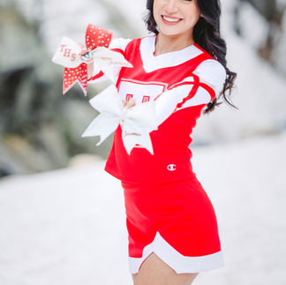 Jasmin smiling warmly while holding two cheer bows, posing in her red cheerleading uniform amidst the snow.