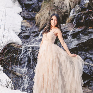 Jasmin standing in front of a snowy waterfall, holding her gown delicately and exuding natural grace.