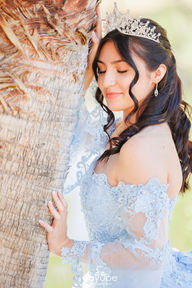 Dezeray in a close-up portrait with her eyes closed, leaning against a palm tree, showcasing her elegant blue dress and tiara.