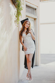 Brenda posing gracefully while leaning on a sunlit wall, her graduation cap and gown complementing her elegant white dress and strappy heels.