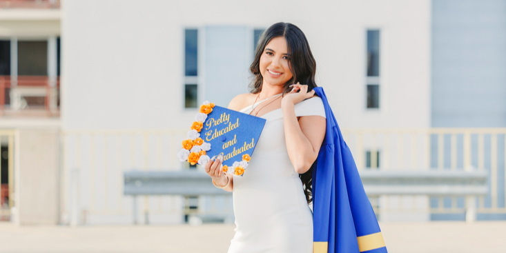 Elegant young graduate Julissa in a white dress draping her blue gown over her shoulder, holding her decorated cap with pride.