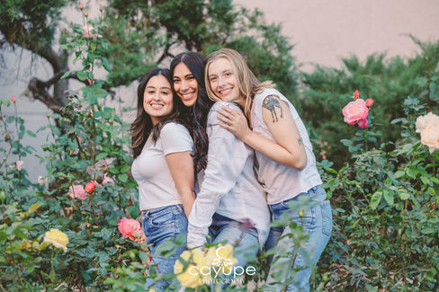 Diana and her two closest friends pose closely together in a rose garden, surrounded by blooming flowers, sharing a beautiful and heartfelt moment.