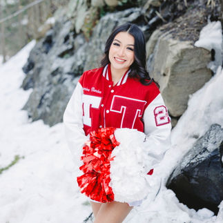 Jasmin sitting on a snowy rock, wearing her cheerleading jacket with her name embroidered on it, holding pom-poms.