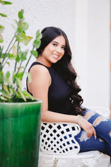 Young woman in a black top and ripped jeans sitting on a white chair, smiling with green plants in the foreground.