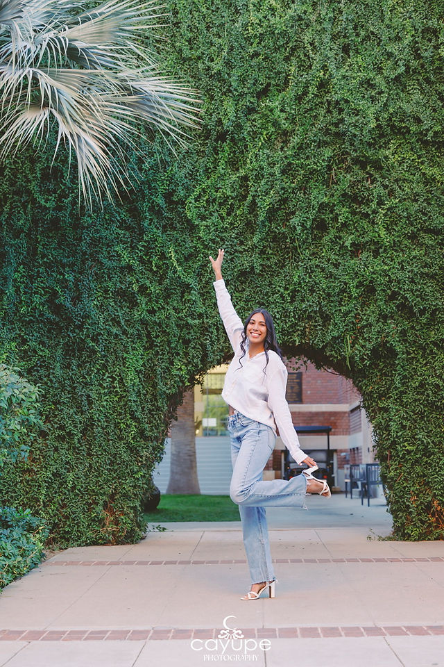 Diana wearing a white blouse and jeans, standing under a large ivy-covered archway on campus.