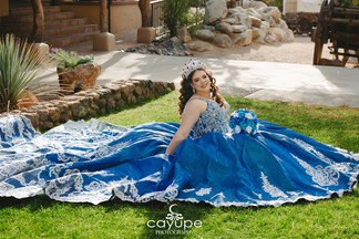Smiling quinceanera portrait of Reina sitting on the grass in her royal blue gown, with rustic ranch details in the background.
