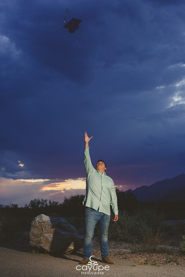 Gerardo tossing his graduation cap into the sky at dusk, with clouds and sunset colors in the background.